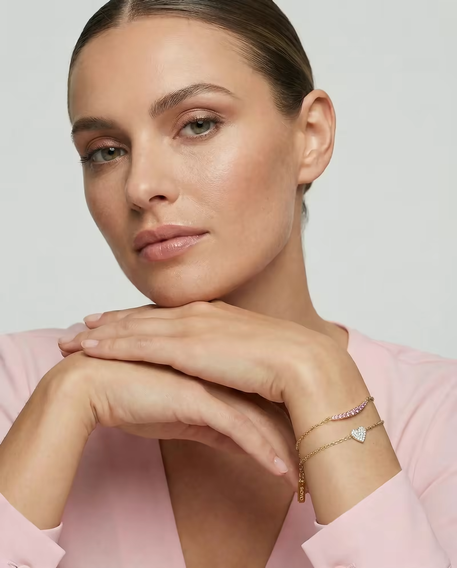 Woman in a pink blazer wearing gold bracelets, hands resting under her chin